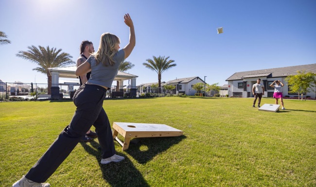 Residents playing a game of cornhole on a large grassy lawn surrounded by palm trees and modern homes under a clear blue sky