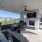 Covered outdoor lounge area with a fireplace, mounted TV, ceiling fan, and cushioned seating, with two residents chatting in a cozy setting.